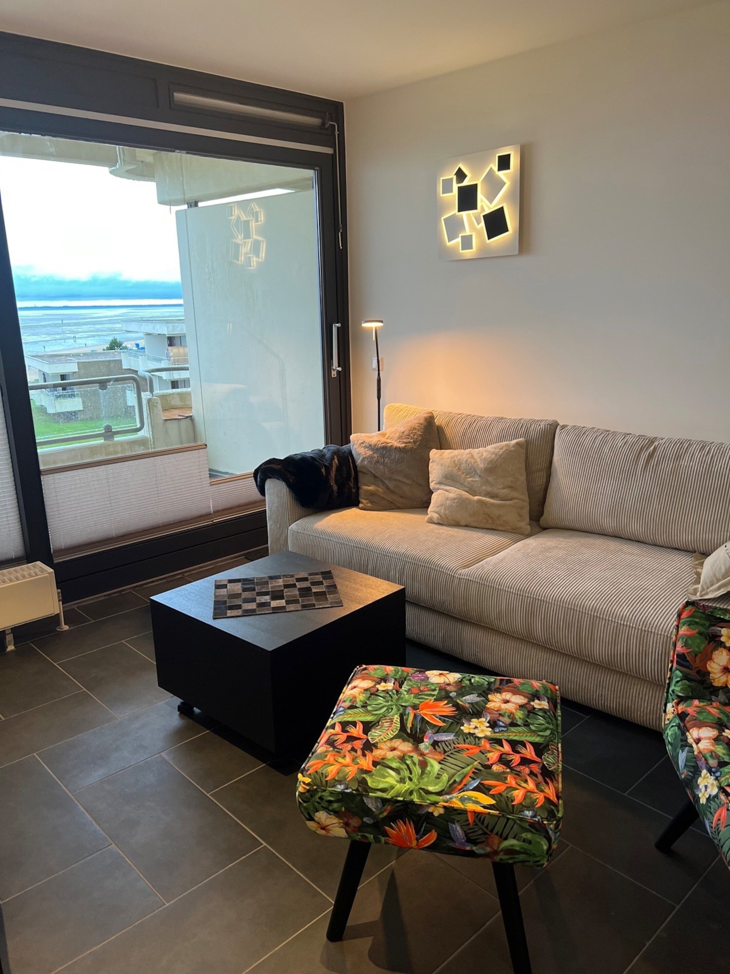 A living room with a sofa, a coffee table, and a patterned ottoman. There is a view of the sea through a glass door.