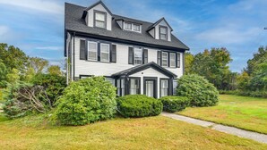 A two-story house with a symmetrical facade, a central entrance with a small porch, and a well-maintained lawn with trimmed bushes. - Charming 2-bedroom apartment in nice Sharon with WiFi, AC near Gillette Stadium  (Sharon)