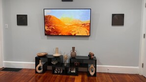 A living room with a mounted TV, black console table with decorative items, and two wall-mounted speakers. - Charming 2-bedroom apartment in nice Sharon with WiFi, AC near Gillette Stadium  (Sharon)