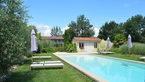 A swimming pool with lounge chairs and umbrellas, surrounded by greenery and a building with a tiled roof.