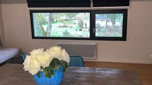 A room with a dining table, a blue vase with white flowers, a window with a view of greenery, and a blue chair.