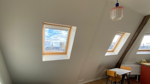 A modern loft bedroom with a sloped ceiling, skylights, a teal sofa, a dining table with chairs, and a kitchenette. - Studio Victoria - Centre de Paris (Paris)