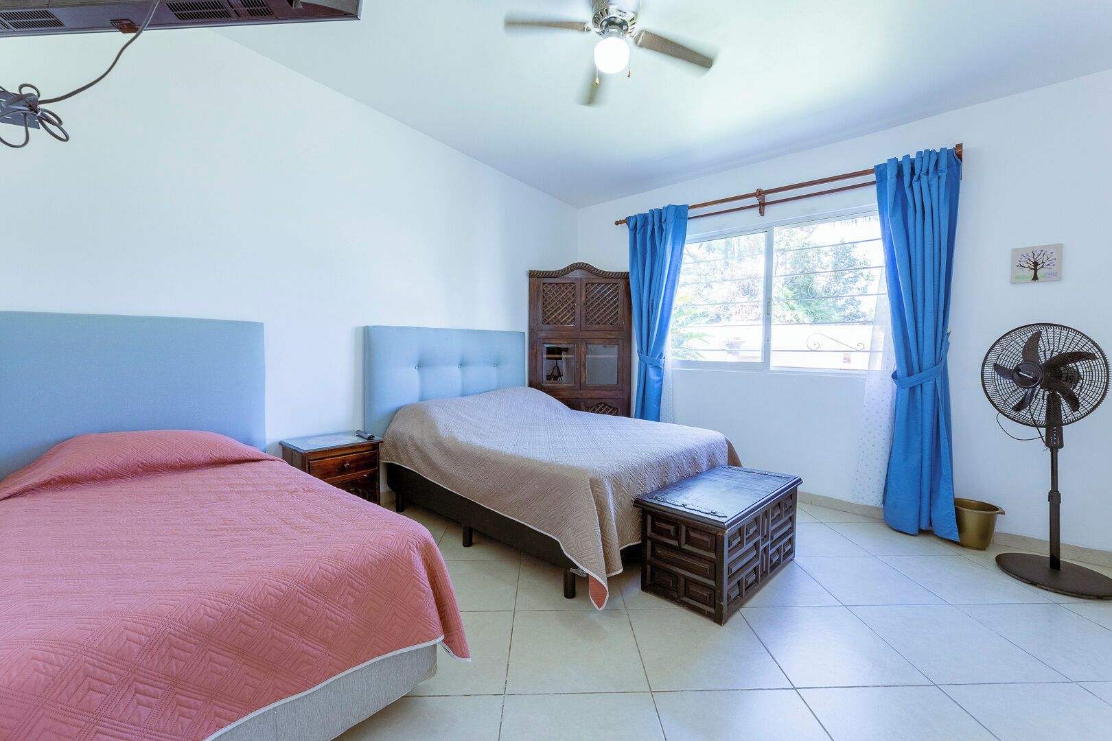 A bedroom with two beds, a nightstand, a wooden cabinet, a standing fan, and a window with blue curtains.