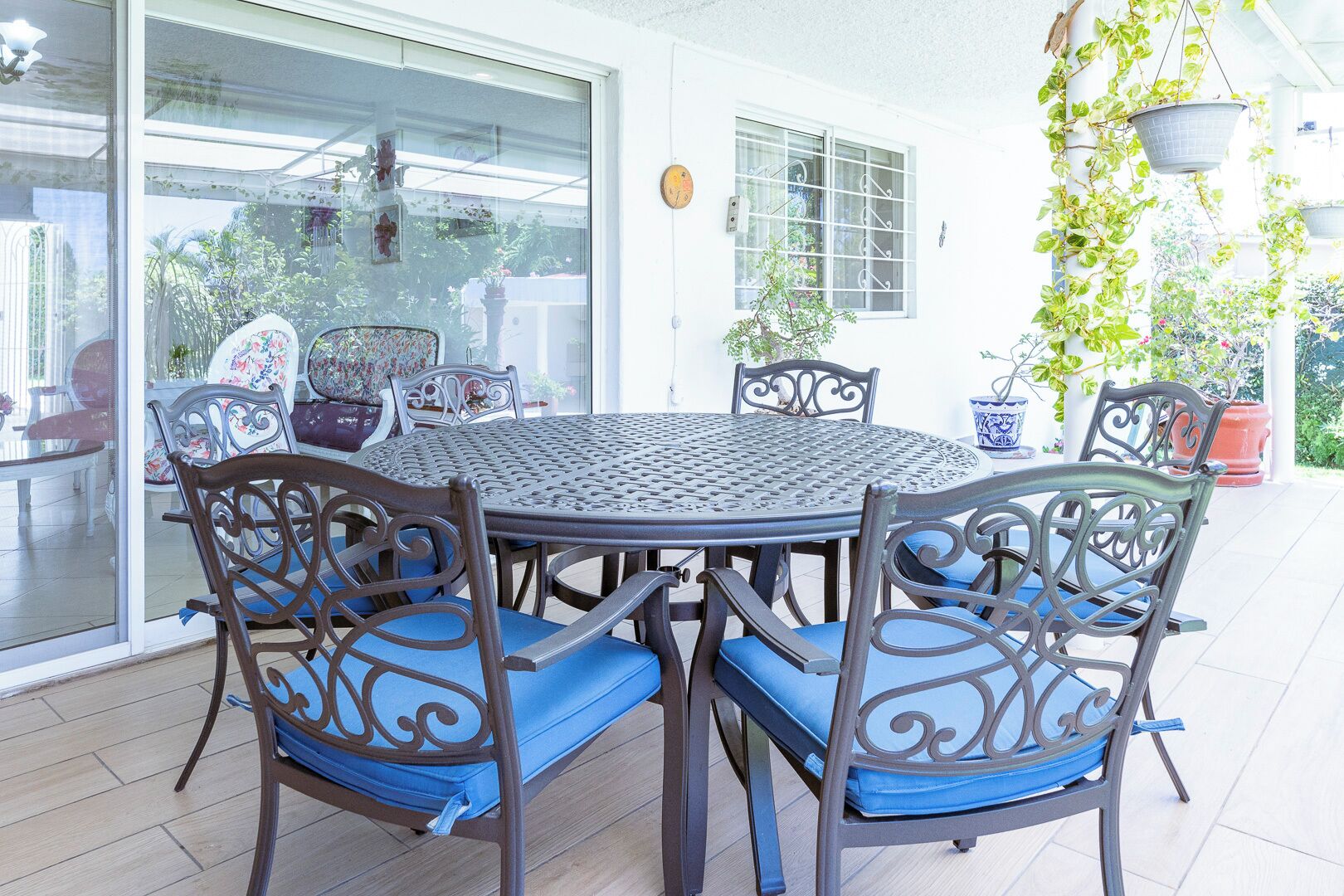 An outdoor patio with a round metal table and chairs, surrounded by potted plants and a glass door leading to a garden.