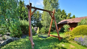 Un’altalena di legno in un giardino, con un’altalena in metallo, circondata dal verde e da una casa con tetto rosso.