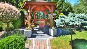 Un gazebo in legno con tavolo e sedie, immerso in un giardino ricco di piante e alberi.