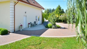 Una casa a un piano con tetto in tegole, vialetto lastricato e un piccolo patio con due sedie e un tavolo.