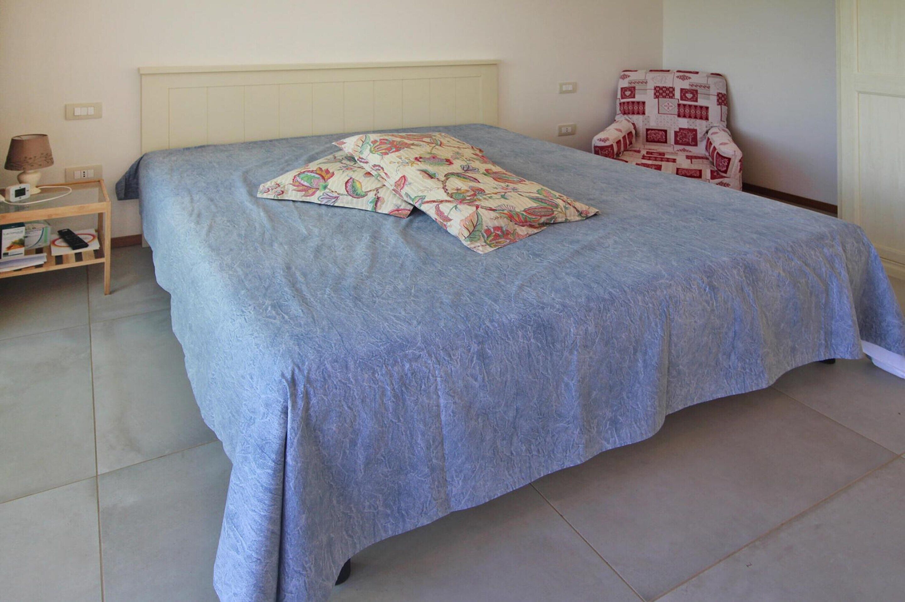 A neatly made bed with a blue bedspread and floral pillows, a bedside table with a lamp, a chair with a patterned cushion, and a tiled floor.