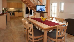 A modern kitchen with wooden cabinets, a dining table with chairs, a TV on a stand, and a staircase. - Apartment on the Spree River in Schlepzig (Schlepzig)