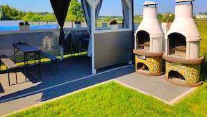 Un patio al aire libre con una zona de estar cubierta, dos hornos de pizza de piedra y una piscina al fondo.