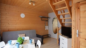 Una habitación acogedora con paredes de madera, un sofá gris, una mesa de comedor con tazas y platos azules, una planta pequeña y un televisor sobre un mueble.