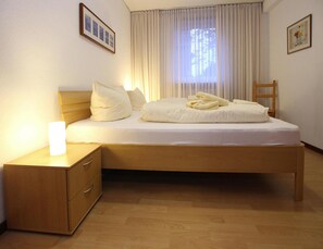 A neatly made bed with white linens and pillows, a wooden nightstand with drawers, a wooden chair, and a window with sheer curtains allowing natural light. - Ferienwohnung in Westerland auf Sylt (Sylt)