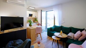 Ein modernes Wohnzimmer mit Flachbildfernseher, einem grünen Sofa, einem kleinen runden Tisch mit einem Getränk und einer Küchenzeile im Hintergrund.