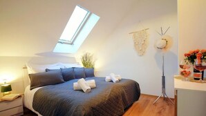 Ein Schlafzimmer mit Dachfenster, einem Bett mit zusammengerollten Handtüchern, einem Nachttisch mit Lampe, einer Stehlampe und einem kleinen Tisch mit Blumen und einer Flasche.
