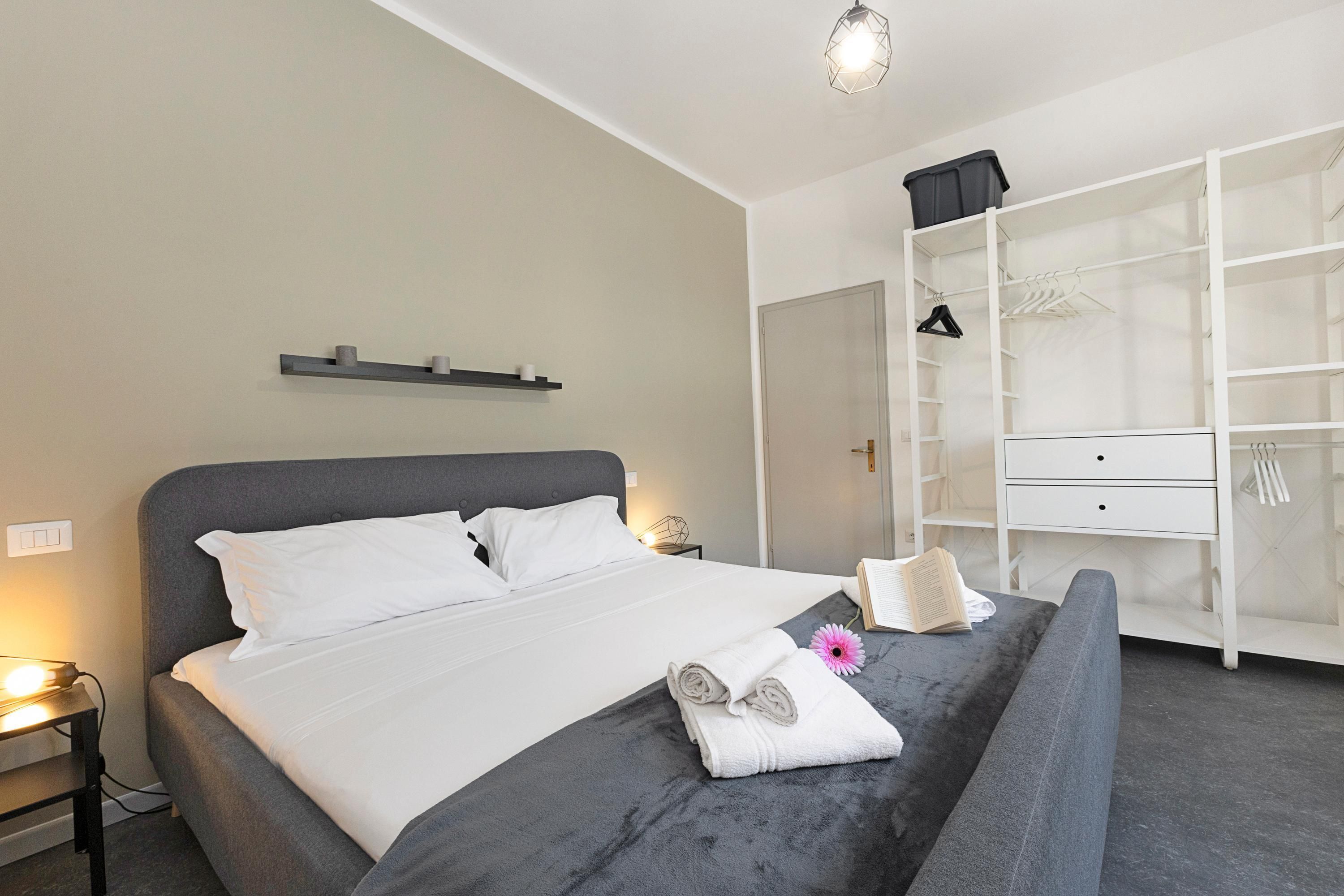 A neatly made bed with white linens, a grey headboard, and a bedside table with a lamp. A large wardrobe with multiple shelves and hanging space is visible to the right.