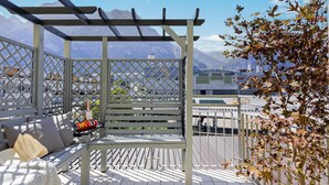 Ein Balkon mit einer Holzbank, einem kleinen Tisch mit Flasche und Gläsern sowie Blick auf Berge im Hintergrund.