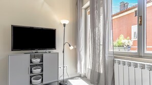 Ein modernes Wohnzimmer mit einem Flachbildfernseher auf einem grauen Schrank, einer Stehlampe und Blick auf ein rotes Gebäude durch transparente Vorhänge.