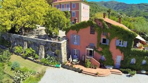 Una villa en la ladera con un muro de piedra, un árbol grande y un edificio rosa con contraventanas azules.