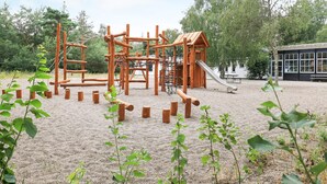 Un'area giochi con strutture in legno da arrampicata e uno scivolo, circondata da sabbia e vegetazione.