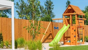 A wooden play structure with a slide and climbing wall, surrounded by a wooden fence and greenery. - Spacious, 2-level Holiday Apartment (Kołobrzeg)