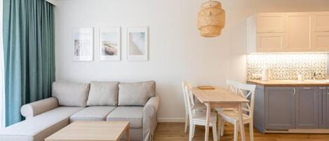 A modern living room with a sofa, a dining table with chairs, a wooden coffee table, and a kitchen area with cabinets and a hanging light fixture.