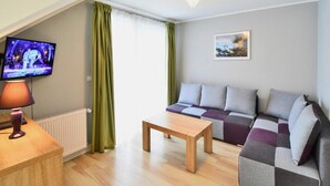 A modern living room with a sofa, a coffee table, a TV, and a chandelier. - Apartment in Darlowko Near Beach & Water Park (Darlowo)