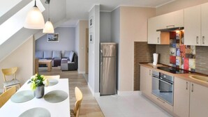 A modern kitchen with a refrigerator, oven, and sink. There is a dining table with chairs and a vase of flowers. A sofa and a chair are visible in the background. - Apartment in Darlowko Near Beach & Water Park (Darlowo)
