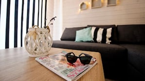 Egy szoba, melyben van egy fa asztal, egy magazin és egy napszemüveg. A szobában egy sötét színű, párnákkal ellátott kanapé és egy fehér fal található, melyre dekoratív fényfüzér van szerelve.