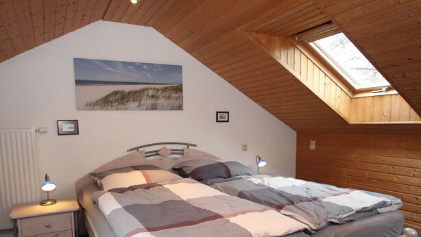 A bedroom with a sloped ceiling, a skylight, a bed with a striped blanket, bedside lamps, and a framed picture on the wall. - Doppelhaushhlfte in Burhave (Butjadingen)