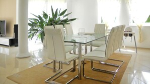 Un espace repas moderne avec une table en verre et des chaises blanches, une plante en pot et un téléviseur en arrière-plan.