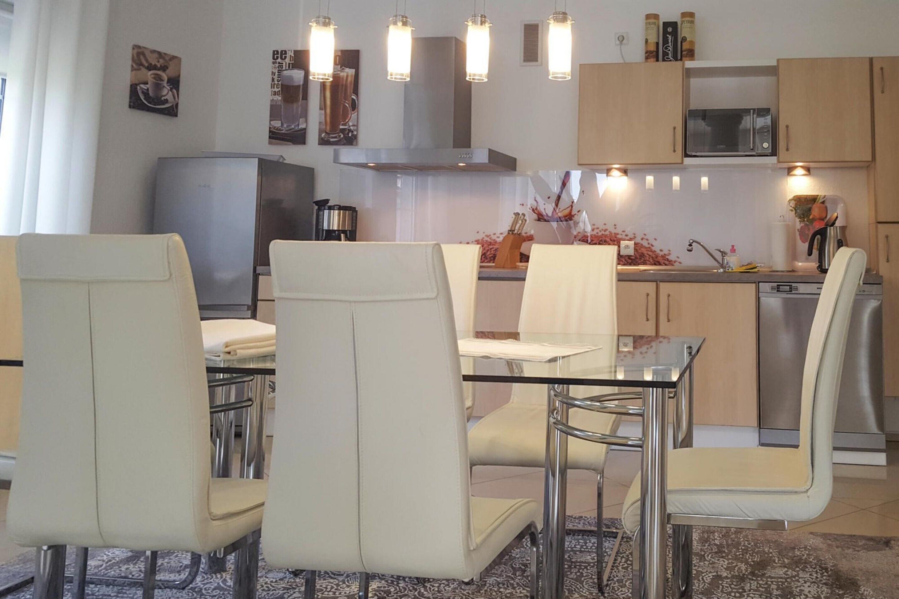 Una cocina moderna con comedor, que incluye una mesa de vidrio, sillas blancas y electrodomésticos de acero inoxidable.