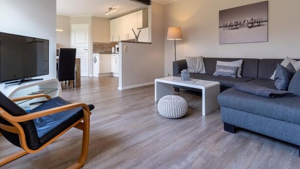 A modern living room with a sofa, a coffee table, a TV, and a dining area with a chair and table. - Ferienwohnung Deichblume (St. Peter-Ording)