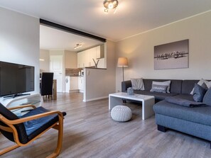 A modern living room with a sofa, a coffee table, a TV, and a dining area with a chair and table. - Ferienwohnung Deichblume (St. Peter-Ording)