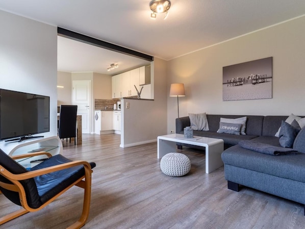 A modern living room with a sofa, a coffee table, a TV, and a dining area with a chair and table. - Ferienwohnung Deichblume (St. Peter-Ording)