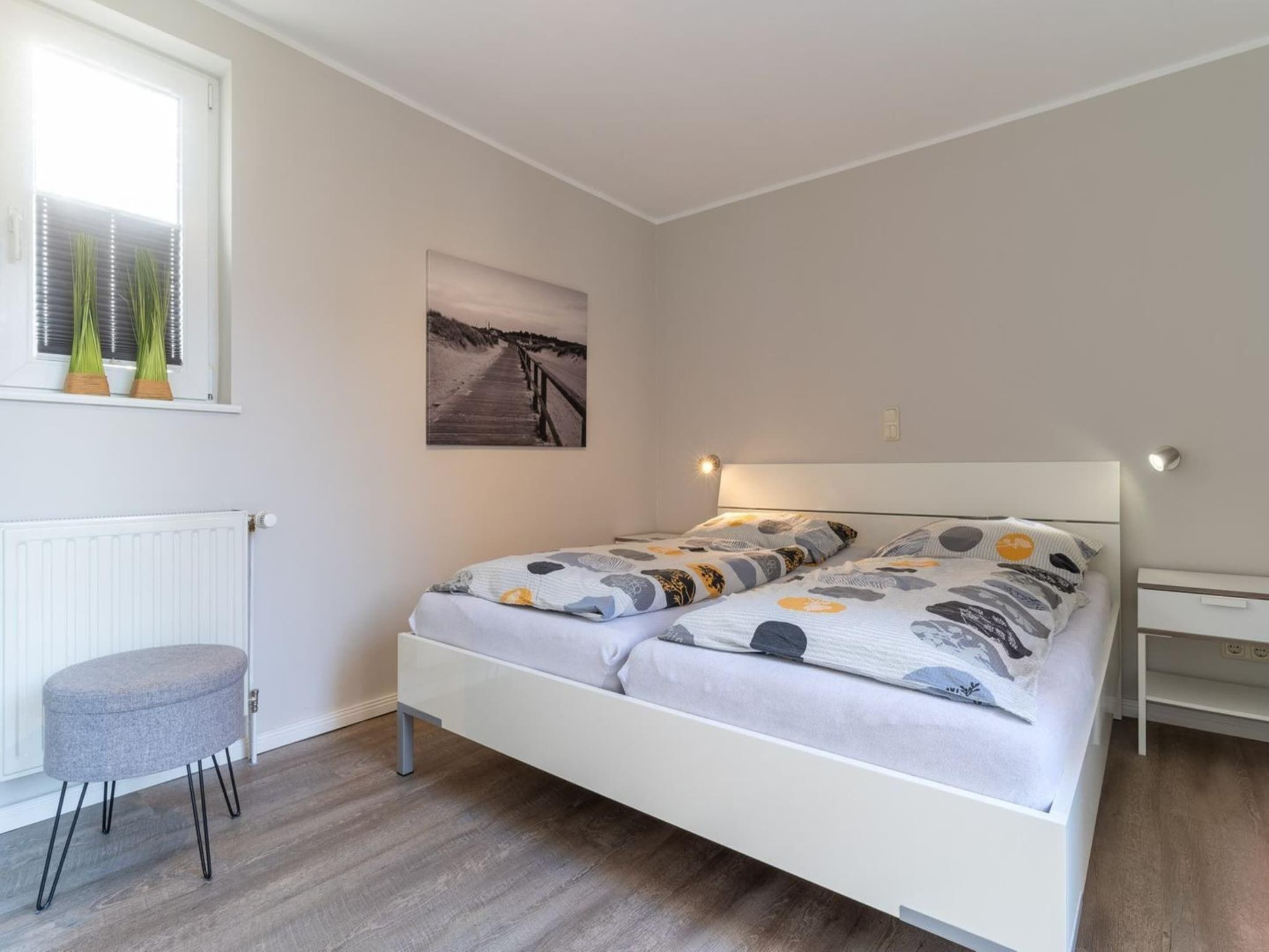 Une chambre à coucher comprenant un lit, une table de chevet, une chaise, un radiateur, une fenêtre avec des stores et un tableau accroché au mur.