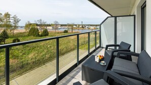 A balcony with a view of a lake and buildings, furnished with wicker furniture and a table with a fruit bowl. - Apartment With a View of the River, Pool and Sauna (Dziwnów)