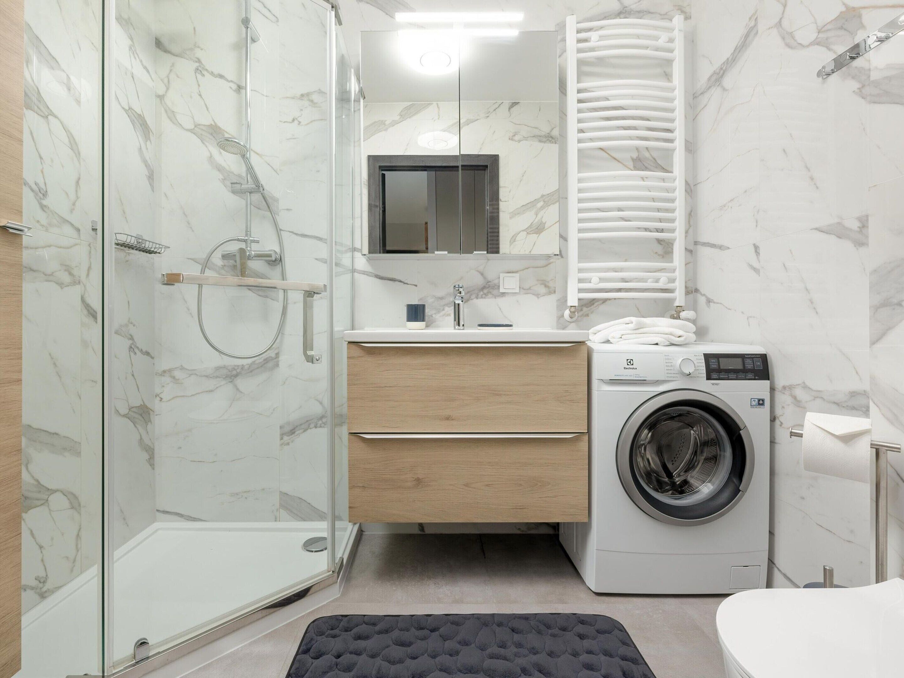 A modern bathroom with a glass shower enclosure, a built-in washing machine, and a wooden vanity.