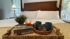 Ein ordentlich bezogenes Bett mit Holz-Kopfstück, einer Nachttischlampe, einem Blumentopf, einem Tablett mit einem Buch, einer Kerze und zwei Tassen.
