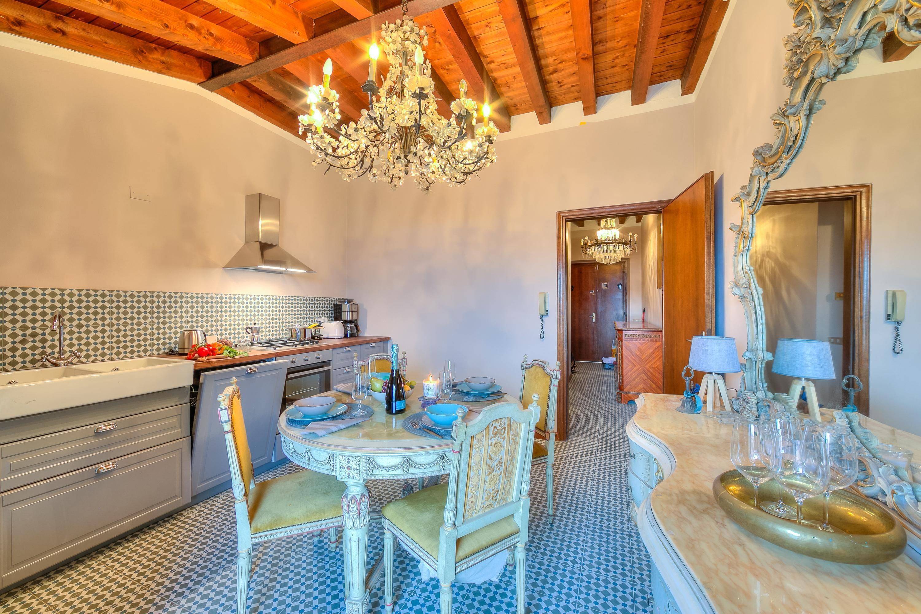 A dining area with a round table, chairs, and a chandelier. There is a kitchen in the background with a tiled backsplash and wooden beams on the ceiling.