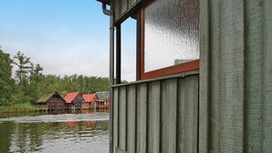 A wooden cabin with a window overlooking a calm lake and several buildings in the background. - Bootshaus, Mirow (Mirow)