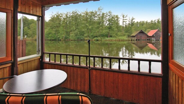 A wooden deck with a table and chair, overlooking a calm lake and a red barn in the distance. - Bootshaus, Mirow (Mirow)