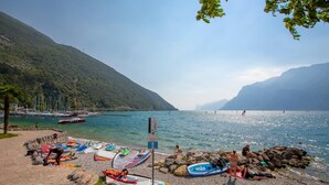 Una playa junto al lago con barcos, orilla rocosa y montañas al fondo.