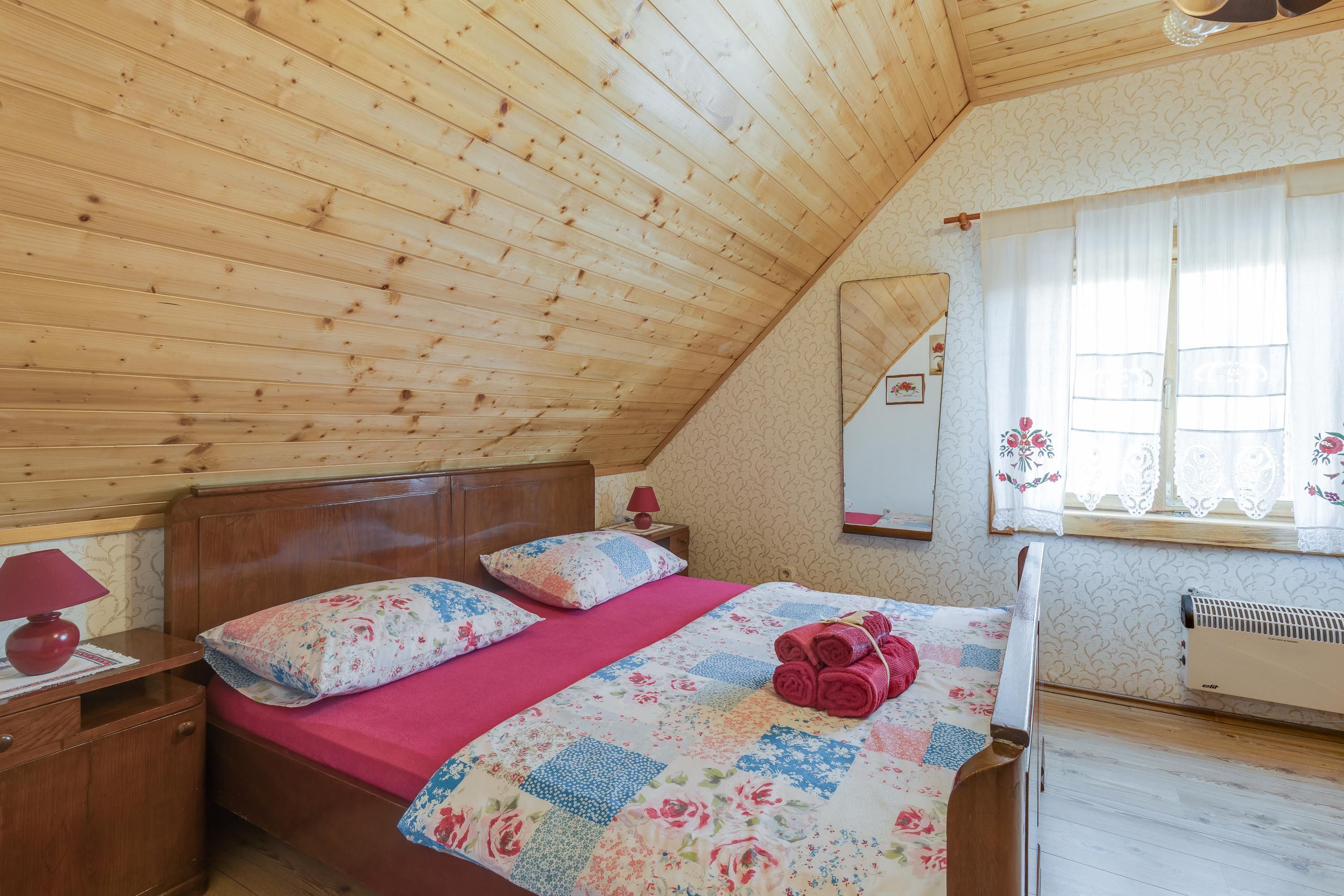 Een slaapkamer met een houten plafond, een bed met bloemenbeddels, een rode lamp en een raam met kantgordijnen.