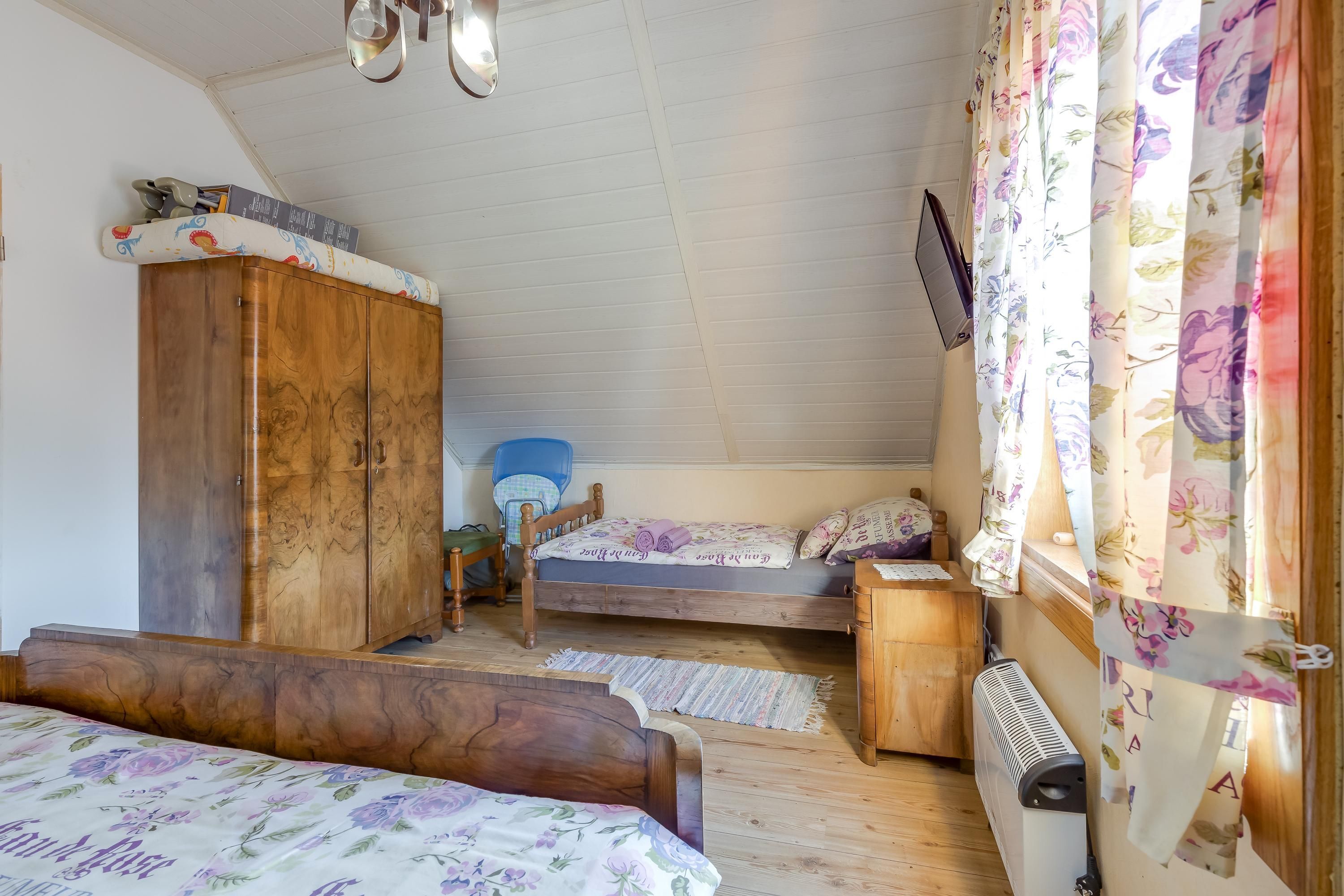Quarto com guarda-roupa de madeira, cama com roupa de cama floral, uma cadeira, uma mesinha e um radiador.