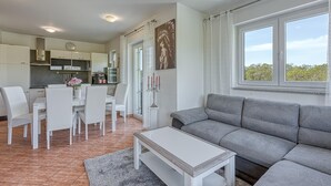 Ein modernes Wohnzimmer mit einer grauen Couch, einem weißen Esstisch und Stühlen, einer Küche im Hintergrund und einem großen Fenster mit Blick.