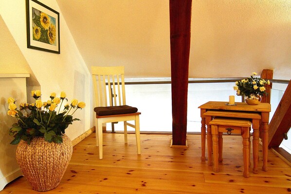 Una silla de madera con cojín, una pequeña mesa de madera con un jarrón de flores amarillas y una cesta de mimbre sobre un piso de madera.