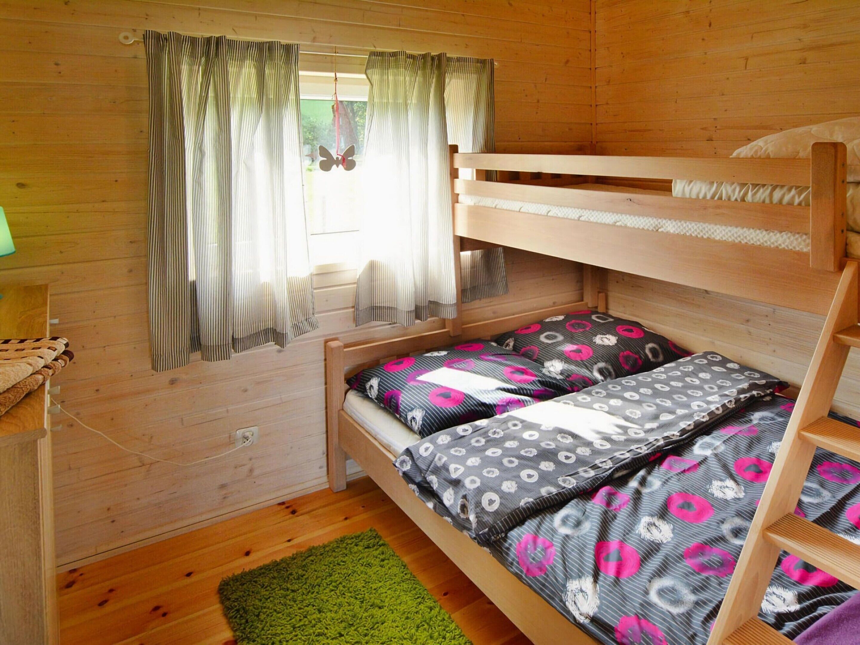 A wooden cabin room with a bunk bed, a window with curtains, and a green rug.