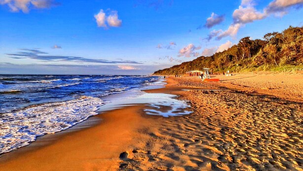 A beach with waves, a lifeguard tower, and a forested hillside. - Holiday Home in Kolczewo Near Baltic Sea (gmina Wolin)
