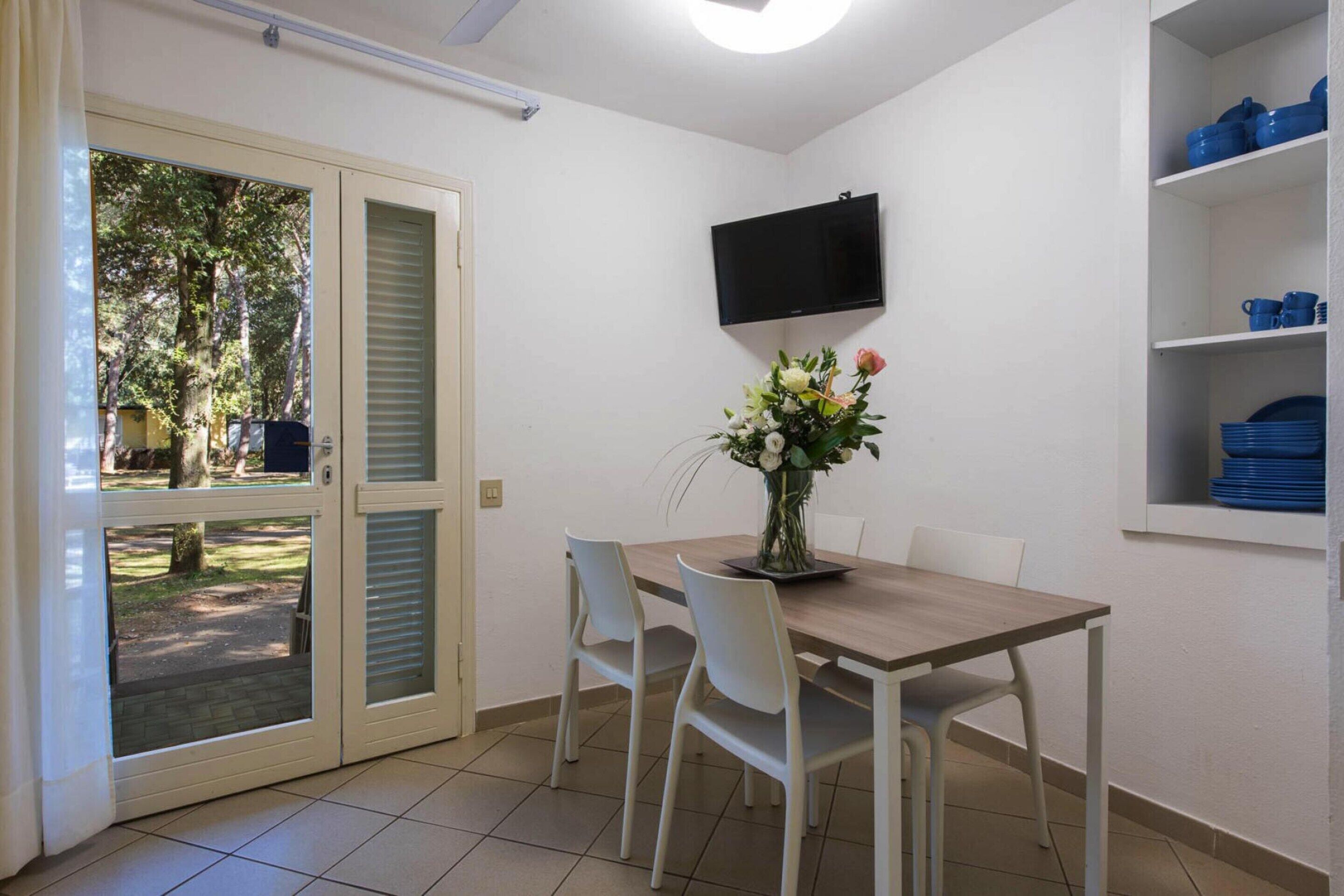 A dining area with a table, chairs, a TV mounted on the wall, and a shelf with blue dishes.