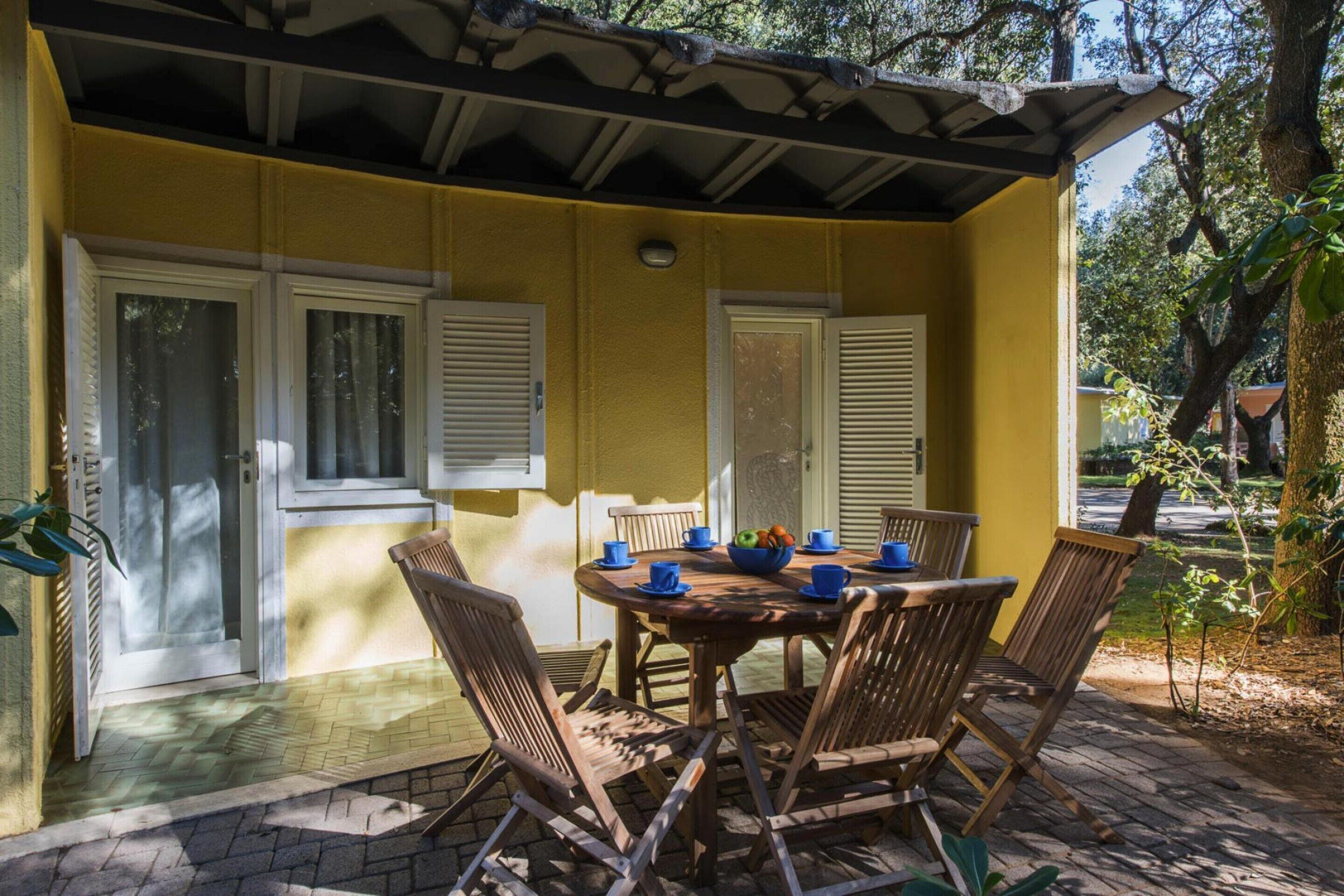 A covered patio with a yellow wall, a round table with chairs, and a bowl of fruit.